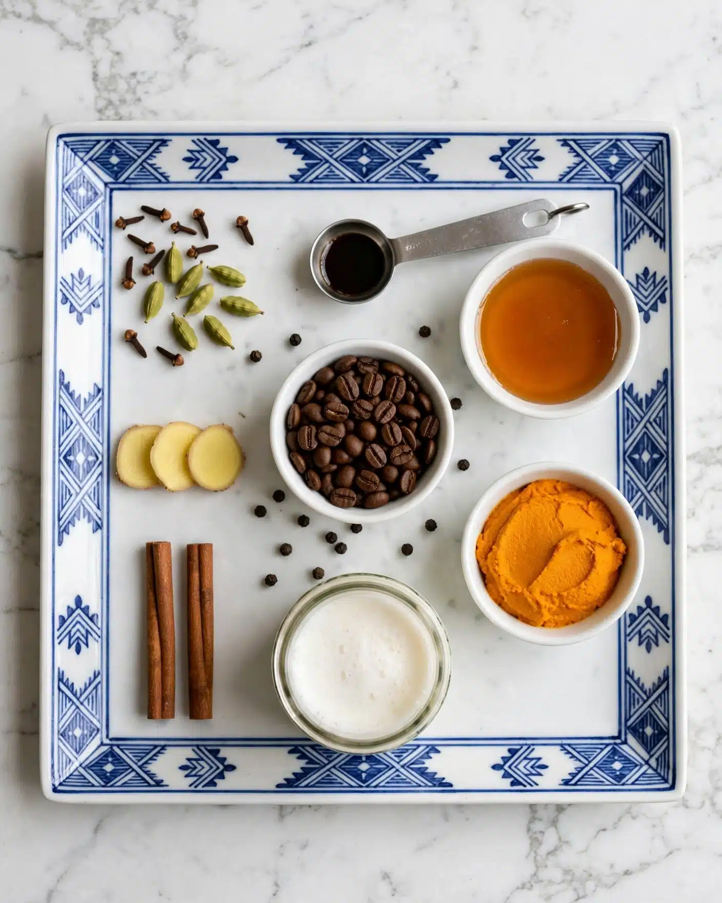 Easy Pumpkin Spice Latte Recipe 4 A top view of a square white plate with blue geometric patterns resting on a white marbled surface. On the plate, several small food items and containers are arranged neatly: at the top left, green cardamom pods and dark cloves scattered; below, three thin slices of pale yellow ginger and two cinnamon sticks placed vertically on the bottom left. In the center, a small white bowl filled with dark brown coffee beans, a measuring spoon with a small amount of dark liquid above it, and scattered black peppercorns nearby. To the right side, a small white bowl with amber-colored honey at the top and below it, a white bowl with bright orange thick paste. At the bottom center, there is a jar filled with white frothy liquid. Photo taken with an iphone --ar 4:5 --v 7