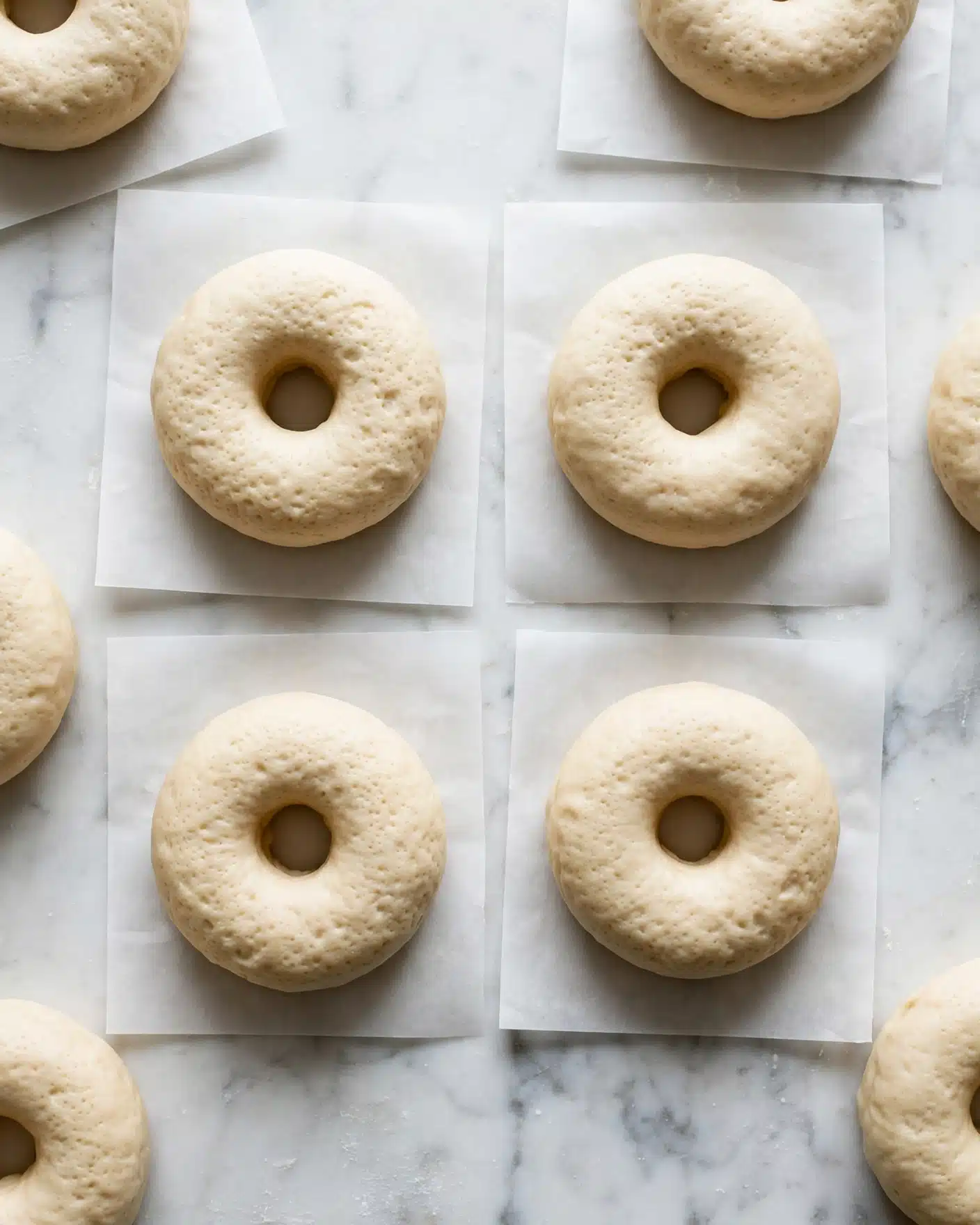 Cinnamon Sugar Doughnuts Recipe 4 The image shows several raw doughnuts arranged on square white parchment papers, placed on a white marbled surface. Each doughnut has one clear round hole in the center and a soft pale beige color with a smooth, slightly dimpled texture. The doughnuts are evenly spaced, with some partly visible at the edges, creating a neat and clean pattern. The lighting is soft and natural, highlighting the dough's light and airy look. Photo taken with an iphone --ar 4:5 --v 7