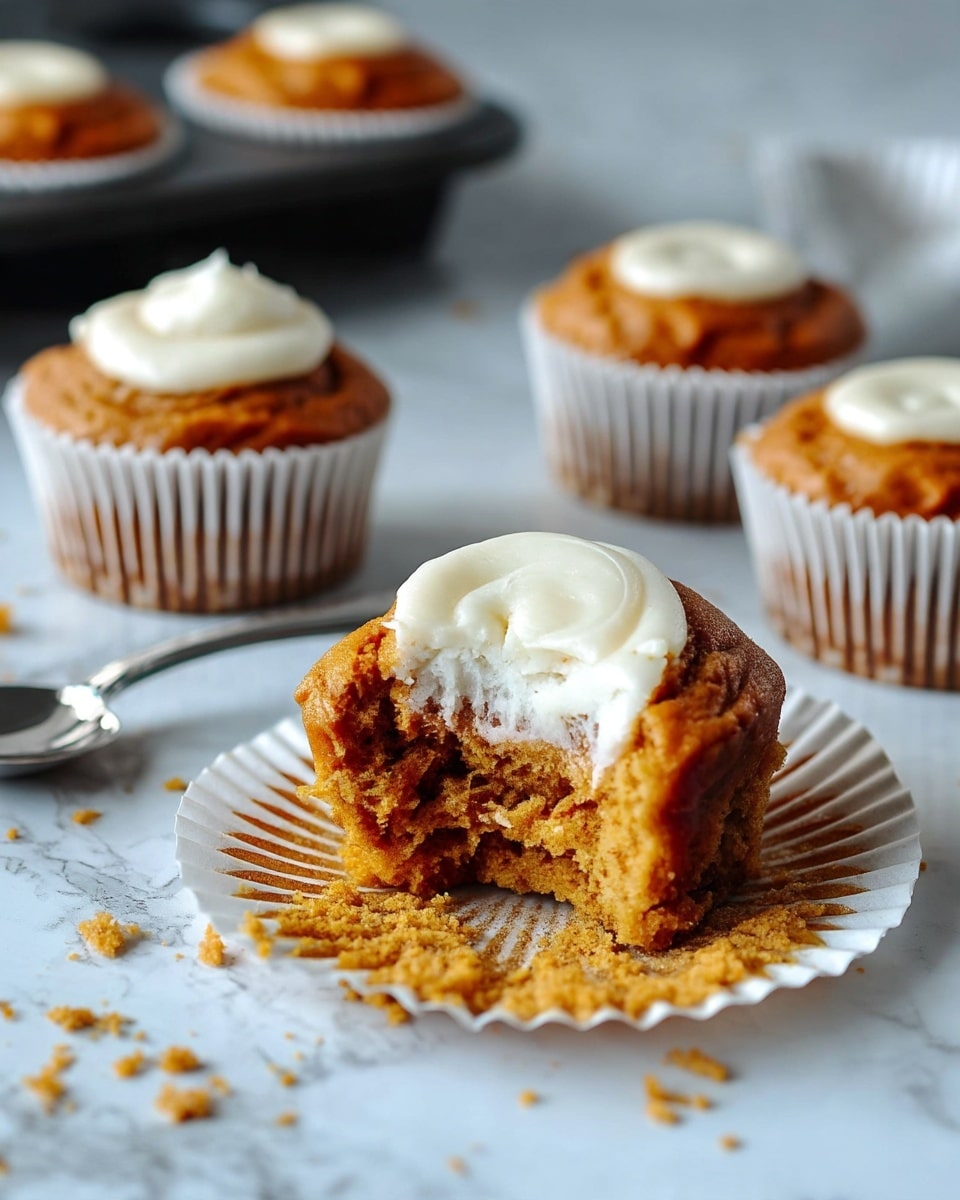 A close-up of an orange-brown cupcake on a white marbled surface with a thick layer of smooth white frosting dolloped on top; this cupcake has a large bite revealing a soft, moist, crumbly inside with white frosting in the center. In the background, there are three more identical cupcakes with white paper liners placed on the white marbled surface, and a dark muffin pan holding more cupcakes with the same frosting can be seen blurred. To the left, some cupcake paper liners are spread with crumbs and a silver spoon resting beside them. Photo taken with an iphone --ar 4:5 --v 7