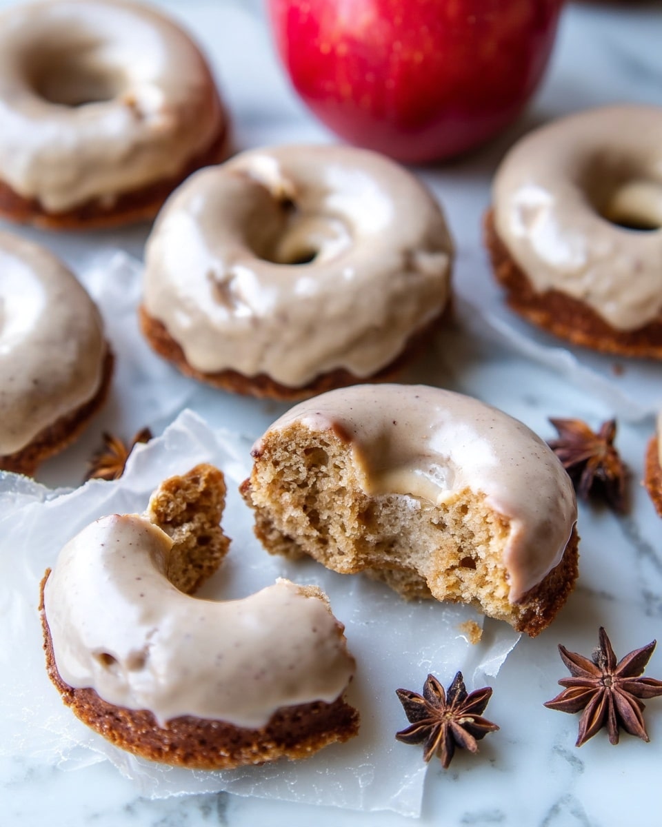 The image shows several donuts with a light brown glaze that looks smooth and slightly shiny, covering the top of each donut. The donuts have one main layer, which is a soft, spongy texture with a warm brown color. Some donuts are whole, while others are broken into pieces, showing the inside with a porous, cake-like crumb. The donuts are placed on a white marbled surface with pieces of white parchment paper under some of them. In the background, there is a red apple, and near the donuts, some brown star anise spices add detail. Photo taken with an iphone --ar 4:5 --v 7