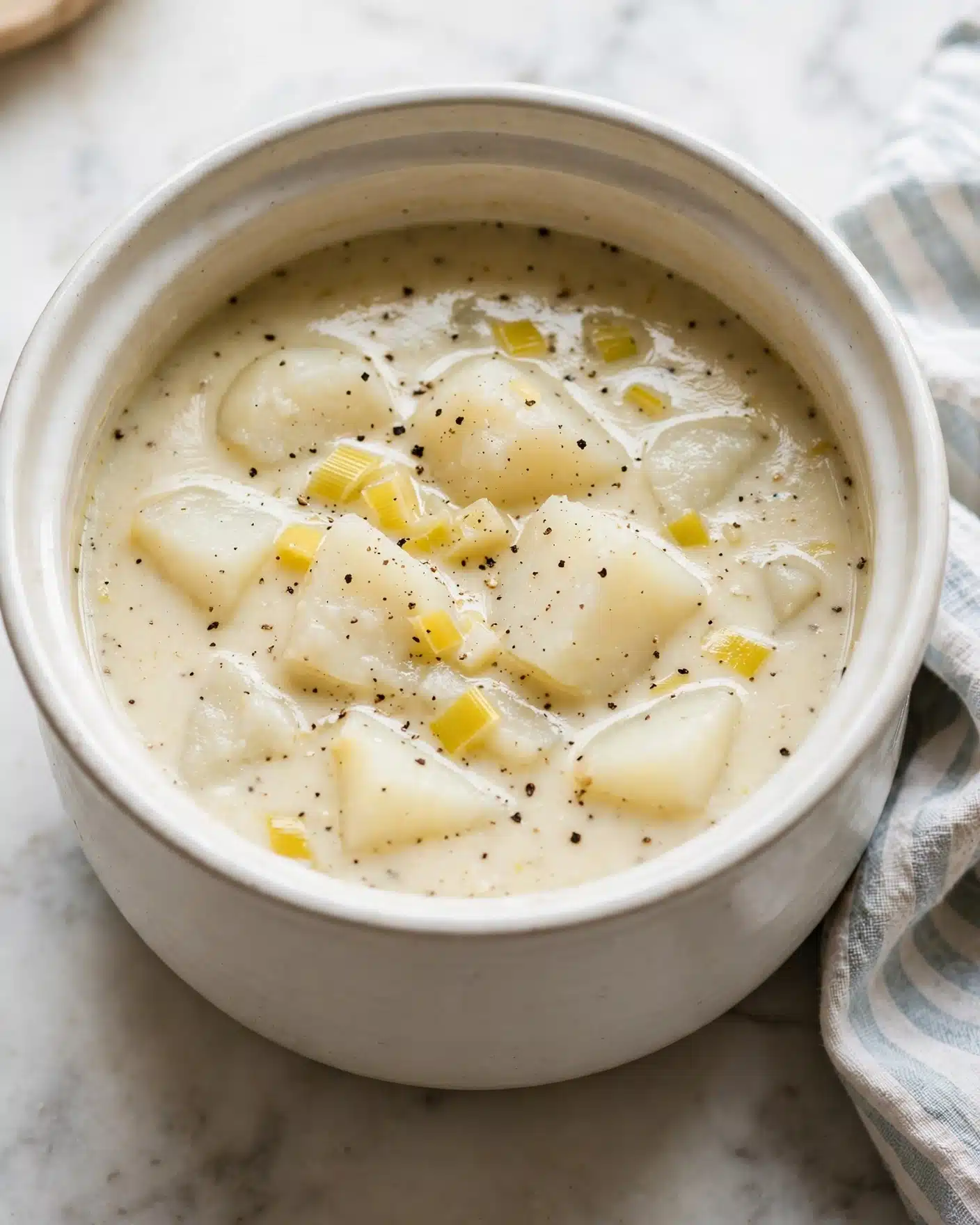 Creamy Potato and Leek Soup Recipe 4 A deep white pot filled with a creamy soup showing two main layers: a thick, pale broth with a slightly shiny texture at the bottom, and large, soft white chunks of potato floating on top mixed with small yellow pieces of leek. The soup surface is seasoned with small black pepper flakes spread evenly. The pot sits on a white marbled surface with a striped cloth partially visible to the side. The overall color tone is light and warm, with the soup looking hearty and gentle in texture photo taken with an iphone --ar 4:5 --v 7