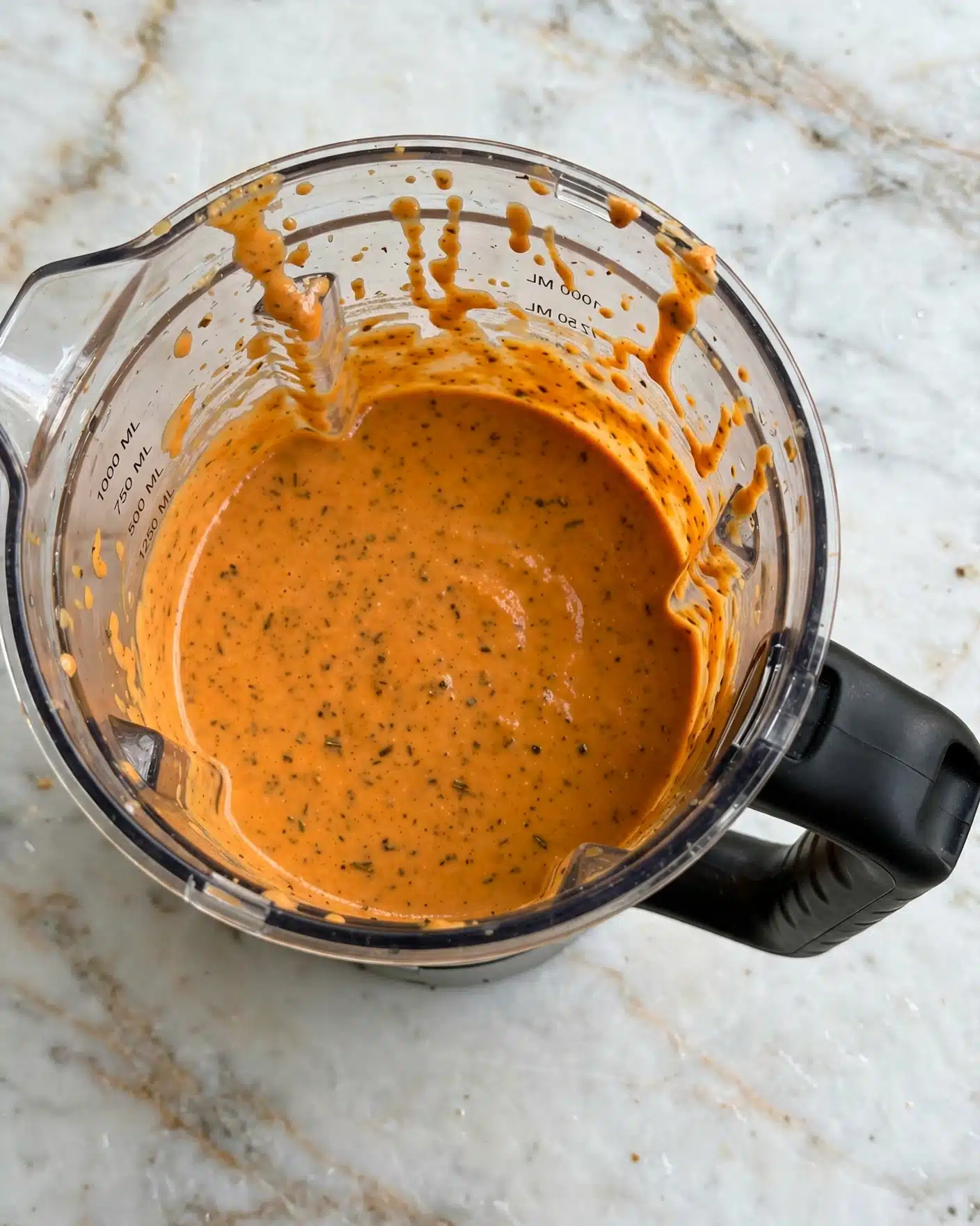 Pumpkin Tomato Soup with Crispy Sage Butter Recipe 4 The image shows a top view of a clear blender jar filled with a smooth, thick orange sauce. The sauce has a slightly shiny, creamy texture with small dark specks spread throughout, likely spices. The blender jar has a black handle on the right side, and it sits on a white marbled surface. The sauce fills about three-quarters of the jar's height, and the jar walls show some sauce residue near the top. photo taken with an iphone --ar 4:5 --v 7