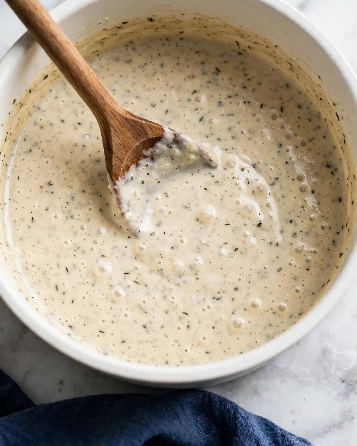 Creamy Potato and Leek Soup Recipe 5 The image shows a close-up of a thick, creamy light beige sauce being stirred by a wooden spoon inside a white pot. The sauce has a smooth, slightly bubbly texture with tiny specks spread throughout. The pot rests on a white marbled surface with a navy blue cloth partially visible at the bottom. Photo taken with an iphone --ar 4:5 --v 7